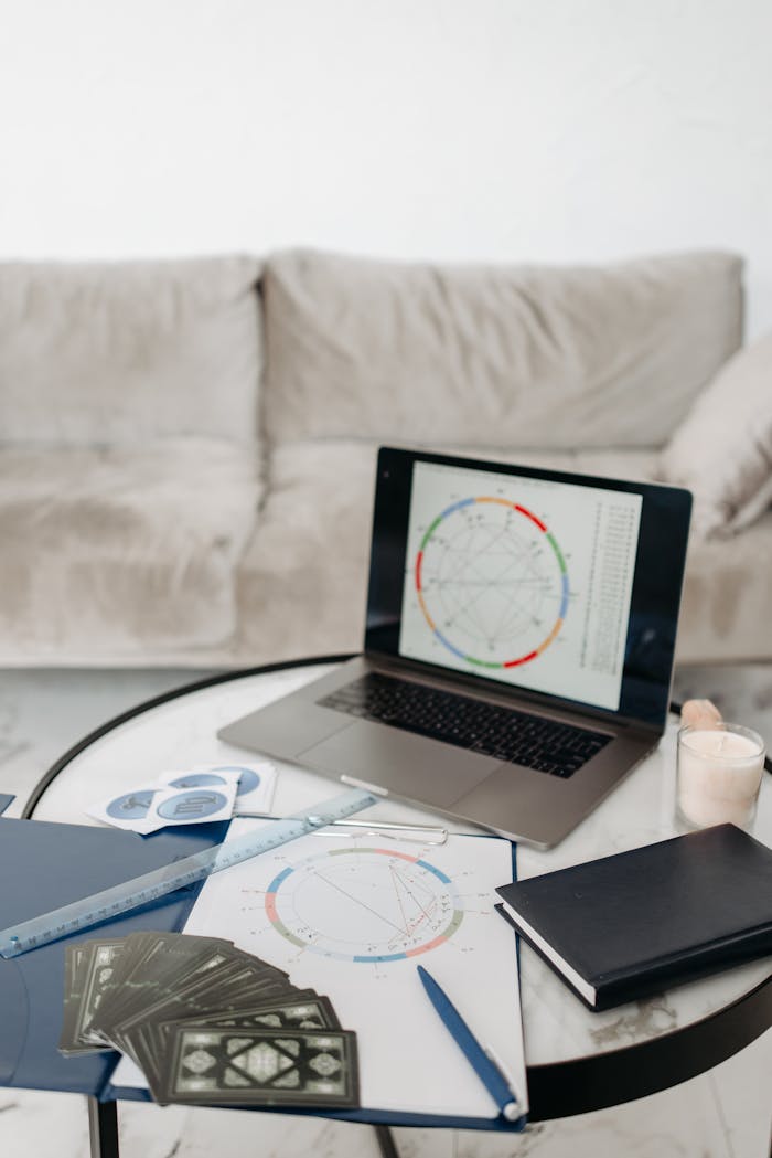 A cozy home office setup featuring a laptop with astrology chart, tarot cards, and a notebook on a table.